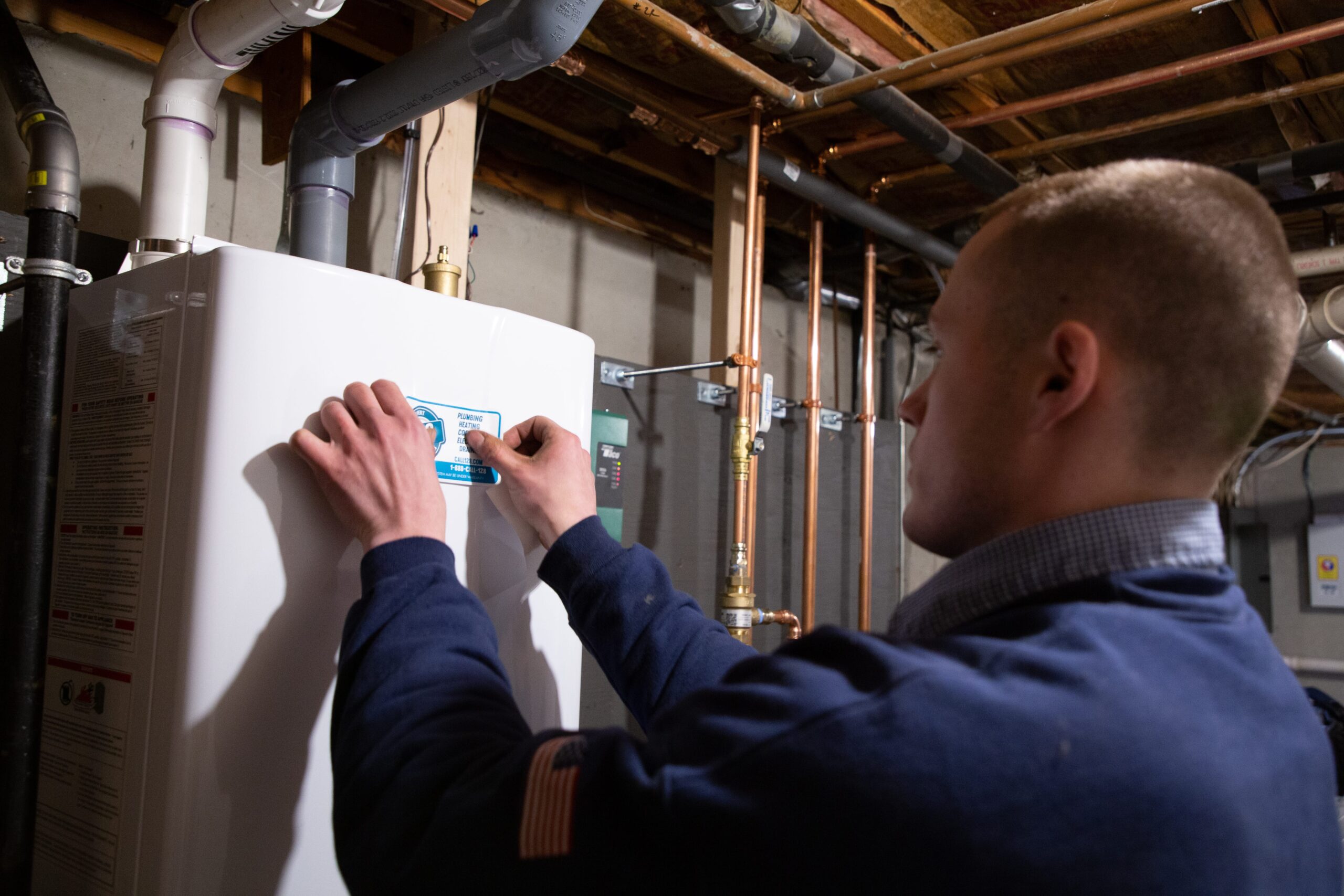 A person in a navy blue uniform applies a maintenance sticker to a white water heater in a basement with exposed pipes and ceiling beams.