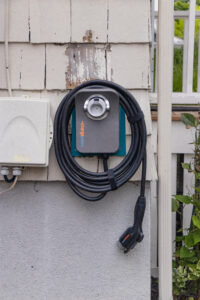A coiled electric vehicle charging cable is mounted on the exterior wall of a house next to a utility box.