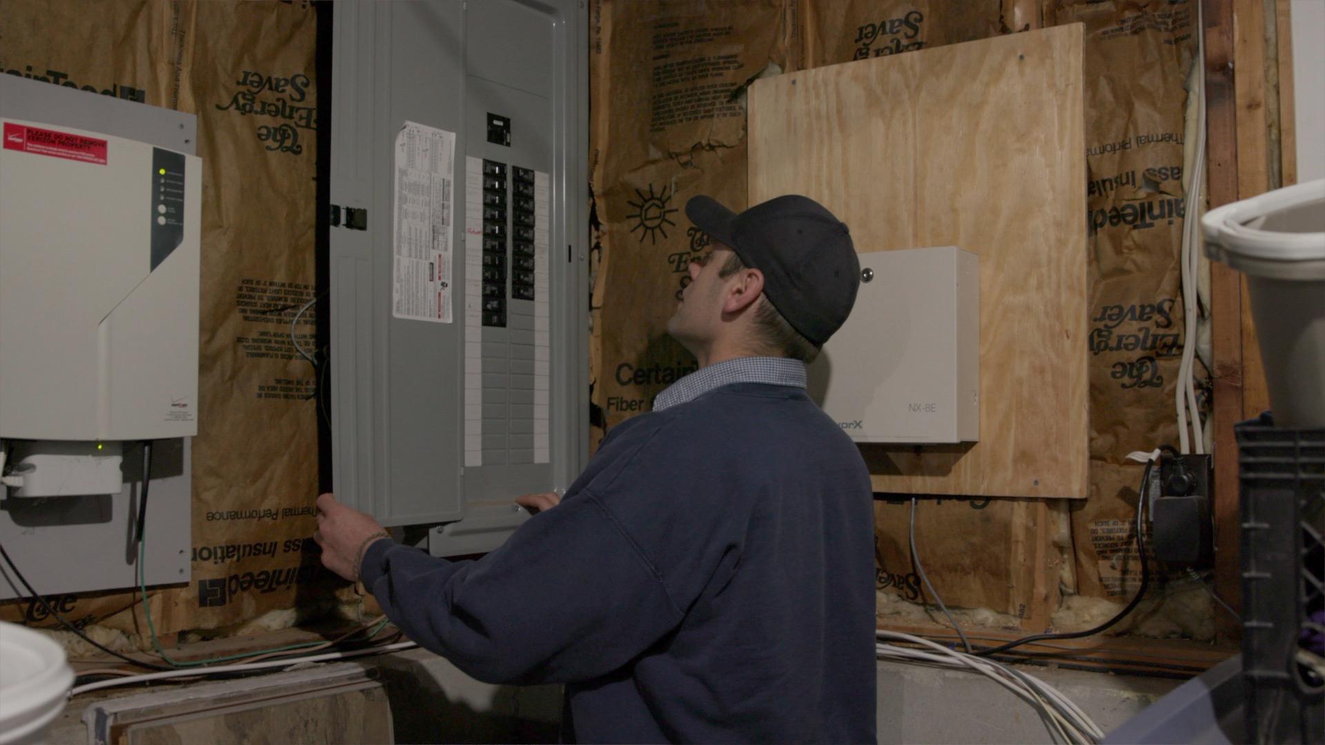 A person wearing a cap and glasses inspects an open electrical panel in a utility room with exposed insulation and wiring.