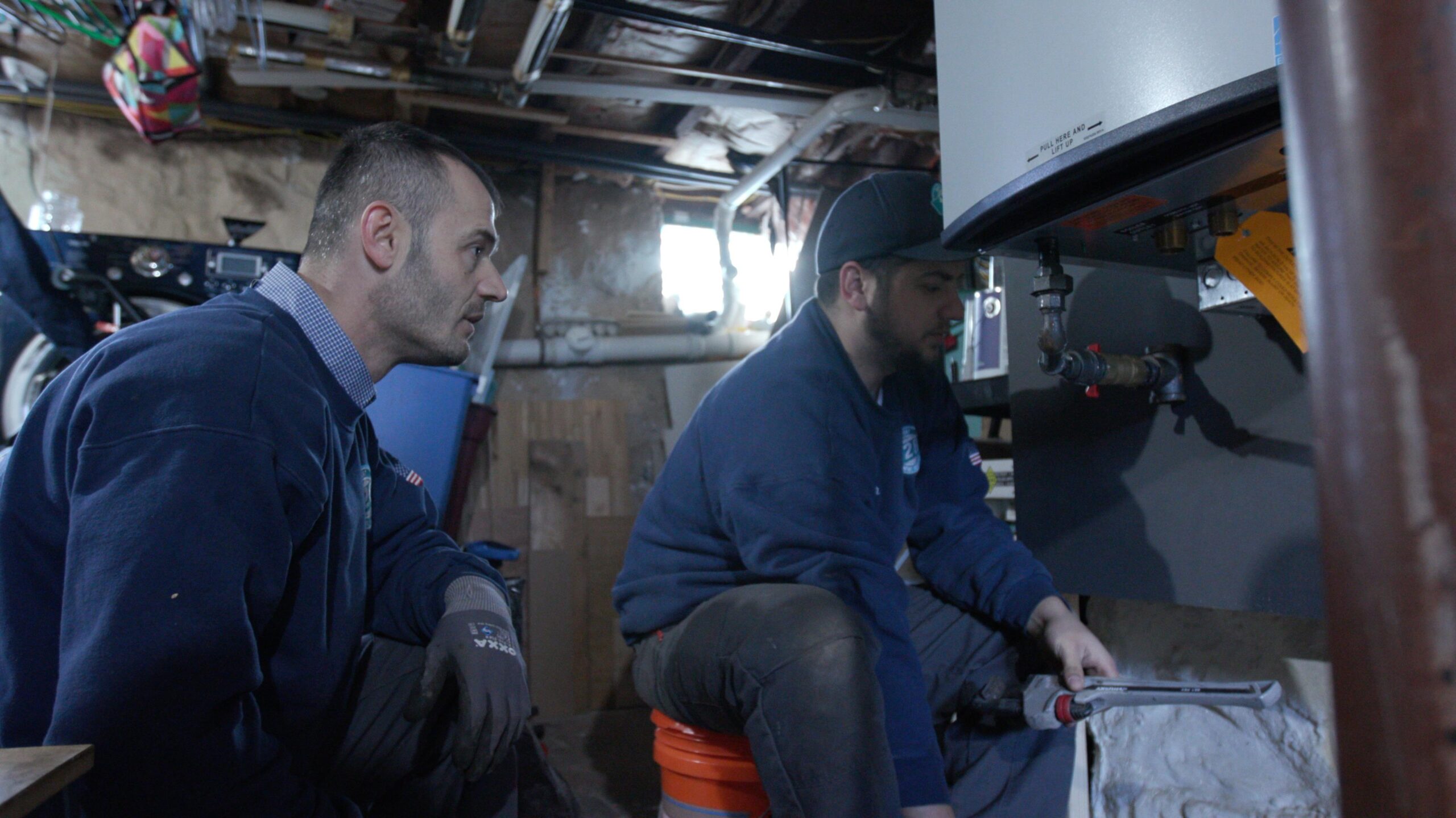 Two workers in blue uniforms examine and adjust equipment in a basement, one holding a wrench and the other observing closely.