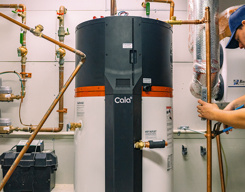 A technician in uniform works on copper pipes next to a large water heater in a utility room with various plumbing equipment.