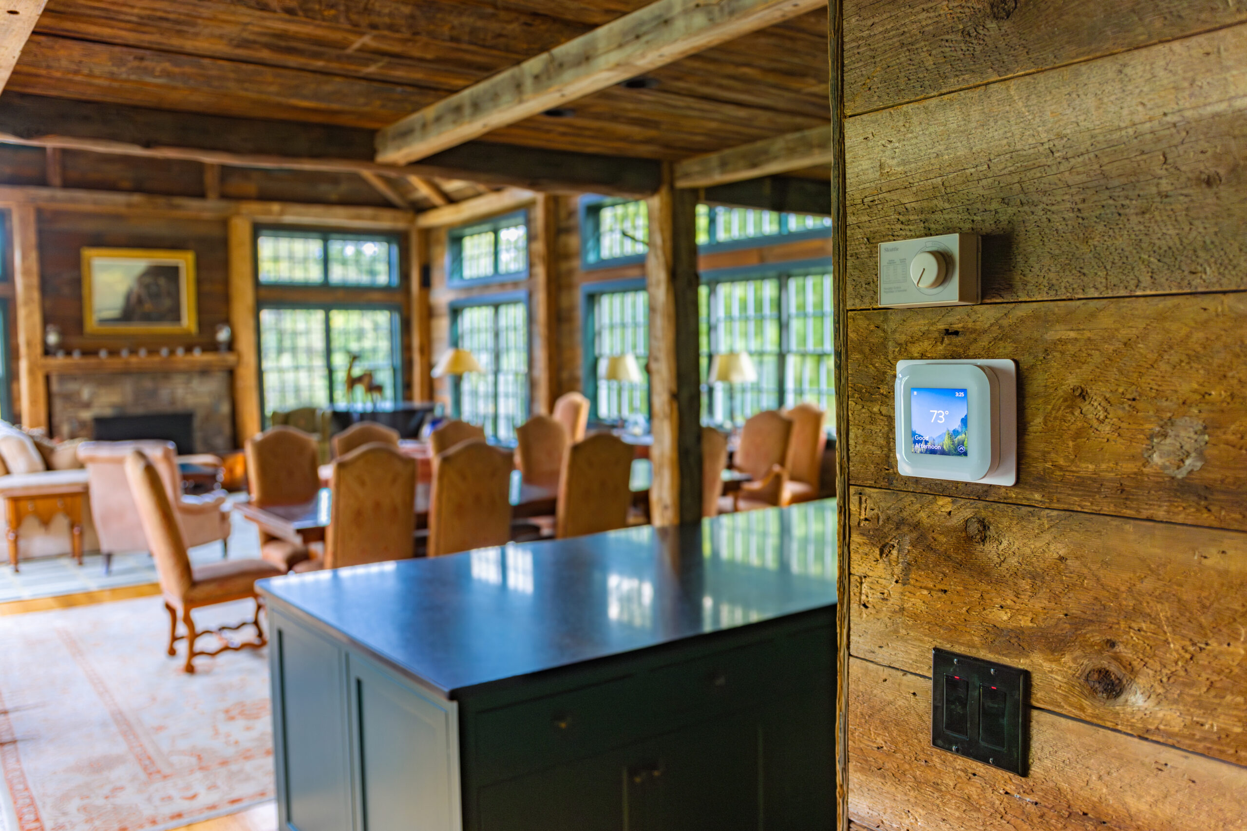 A smart thermostat set to 73°F is mounted on a rustic wooden wall in a sunlit dining and living area with large windows and wooden furniture.