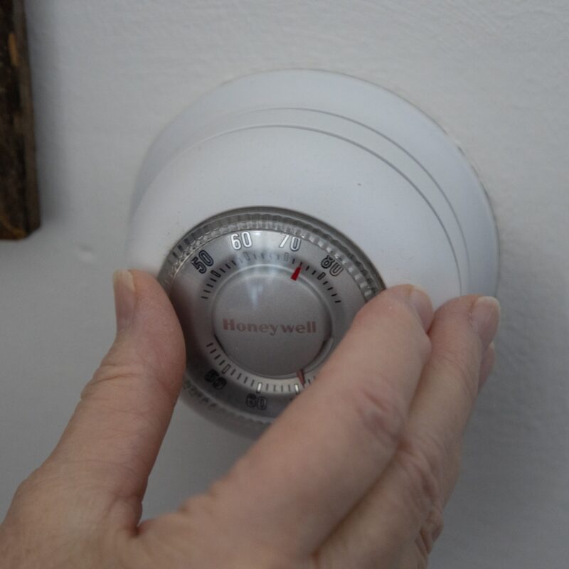 A hand adjusts the temperature on a round Honeywell thermostat mounted on a white wall, reflecting choices like oil vs gas vs electric heat for home comfort.