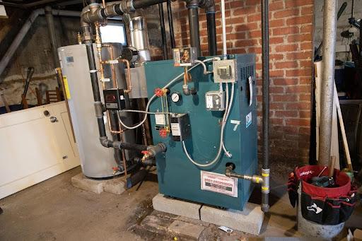 A boiler system with pipes and a water heater tank installed in a basement with brick walls and concrete floor. A red tool bag is on the right.