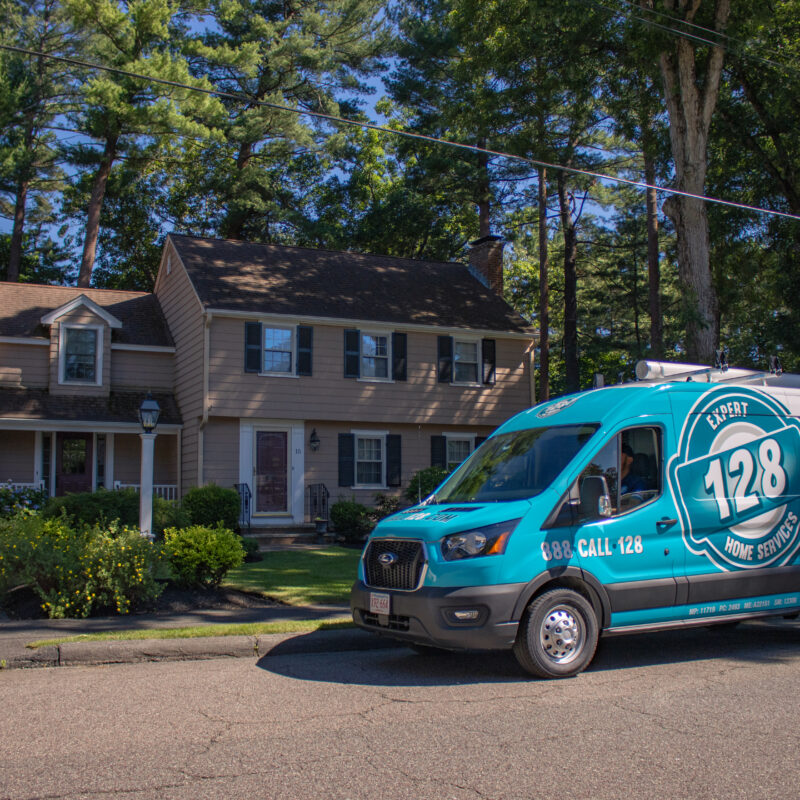 A blue 128 Plumbing, Heating, Cooling & Electric van is parked on the street in front of a two-story suburban house surrounded by trees in Chelmsford, MA.