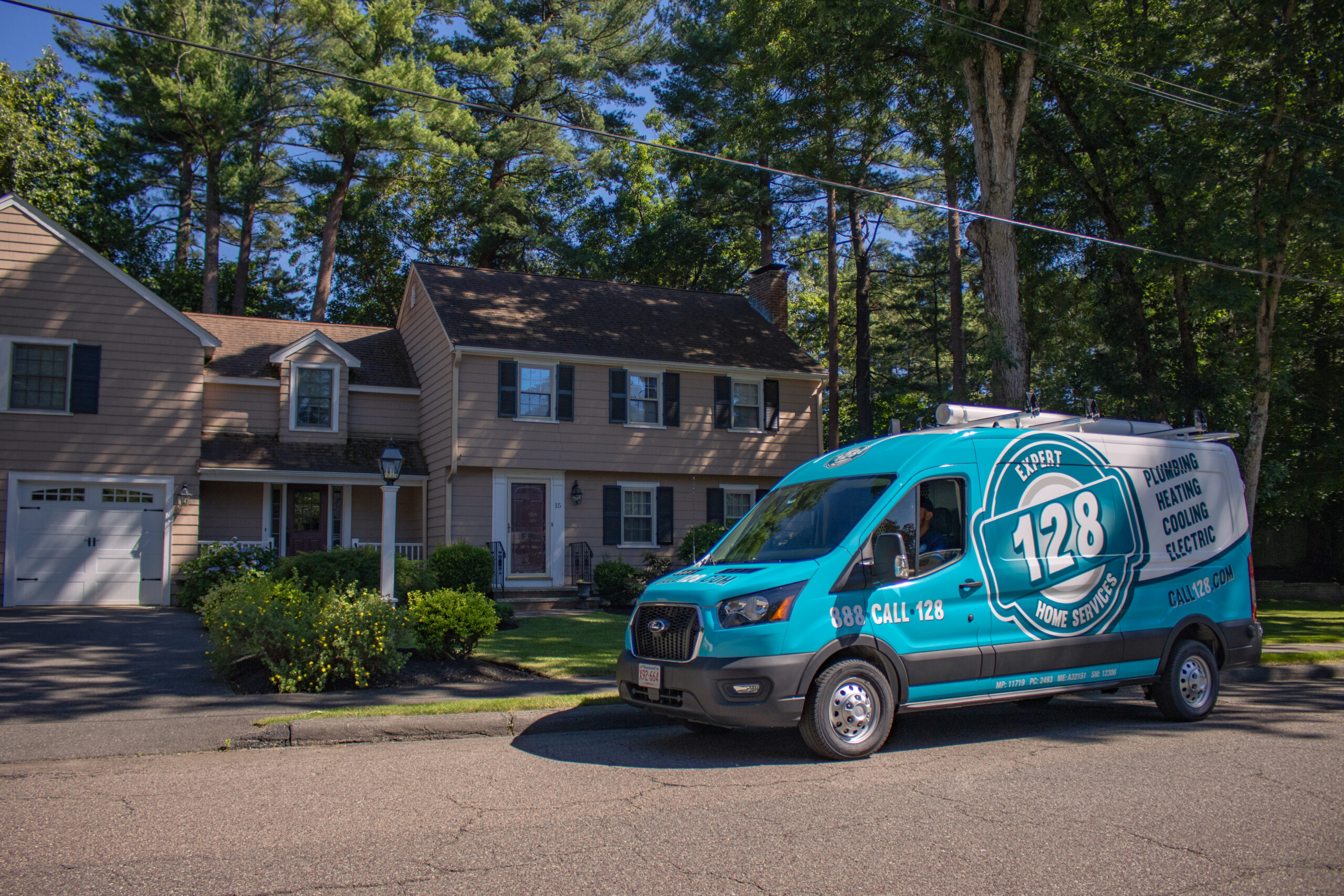 A blue 128 Plumbing, Heating, Cooling & Electric van is parked on the street in front of a two-story suburban house surrounded by trees in Chelmsford, MA.