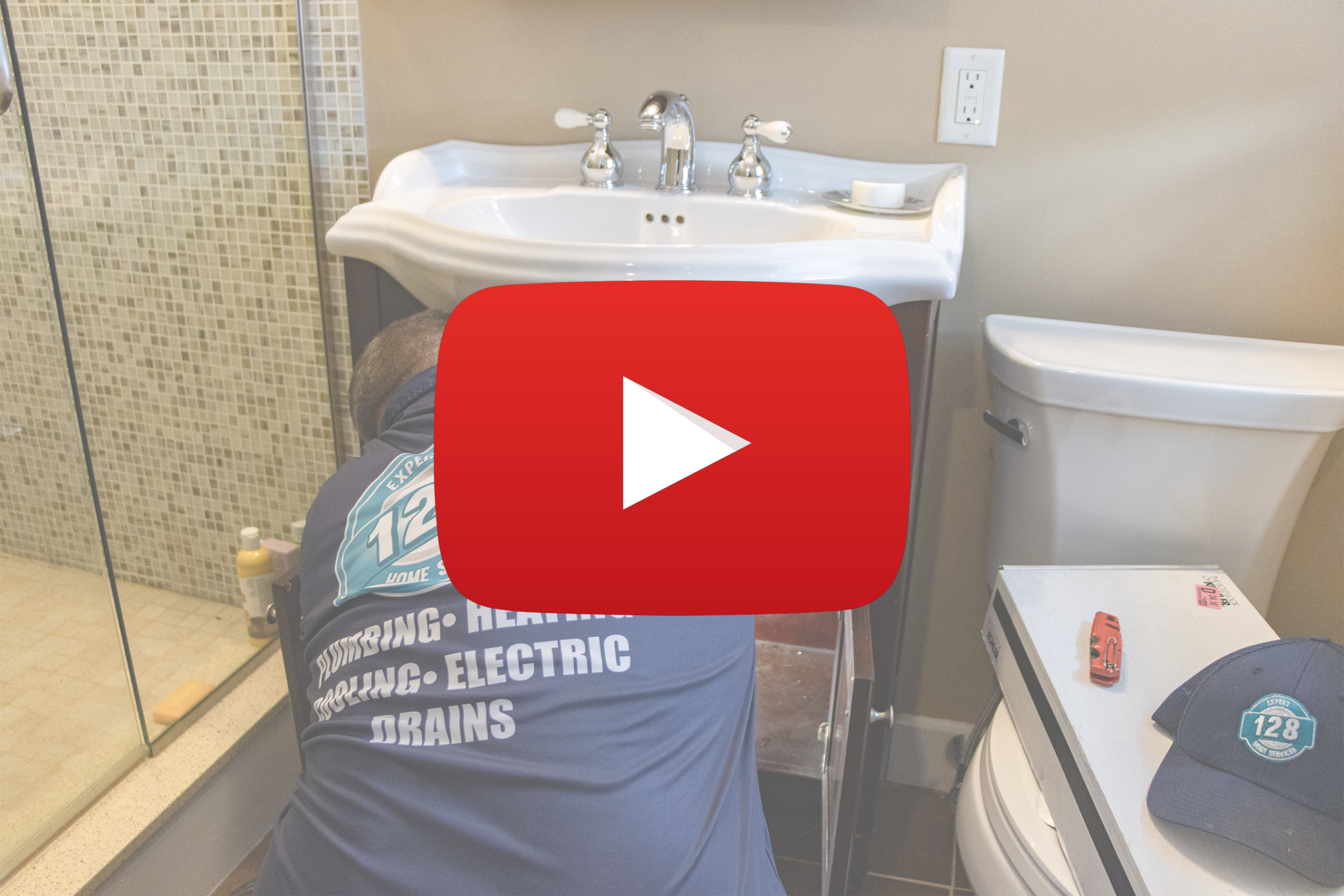 A plumber works on pipes under a bathroom sink next to a toilet, with tools and a hat nearby. A large YouTube play button icon overlays the image.