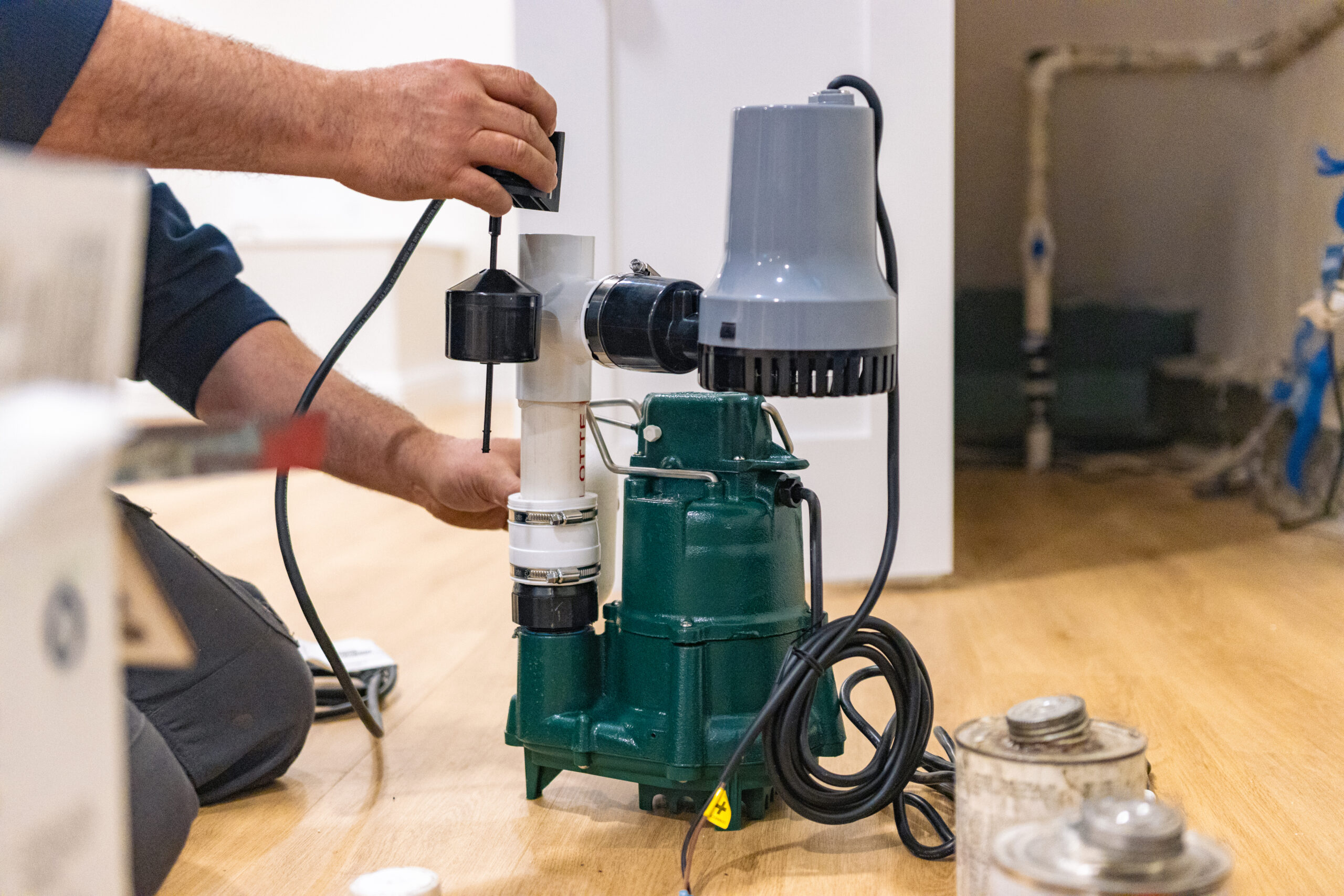 Person installing or assembling a green sump pump with attached float switch on a wooden floor, surrounded by plumbing equipment.