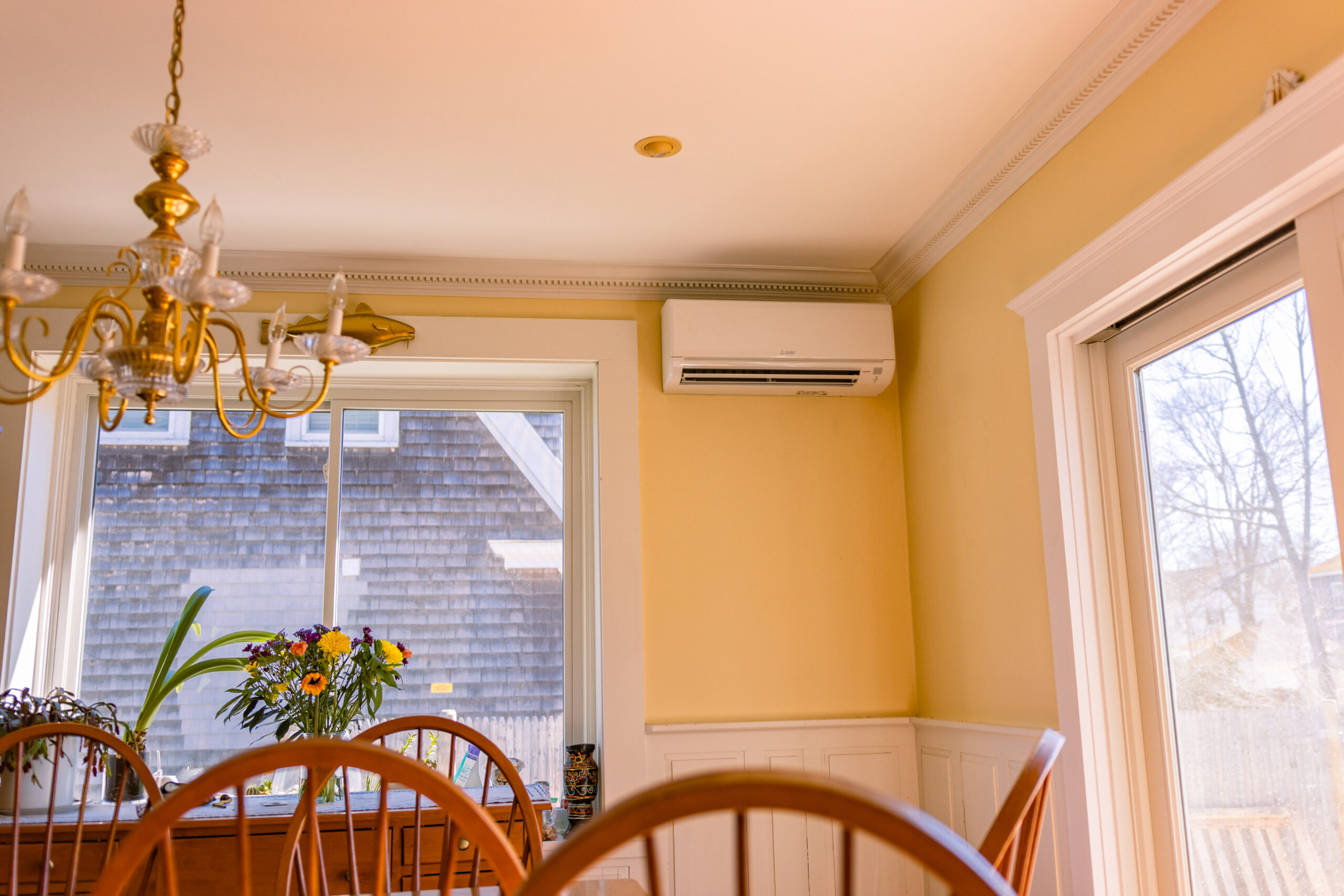 A dining room with wooden chairs, a glass chandelier, a wall-mounted air conditioner, large windows, and a flower arrangement on the table.