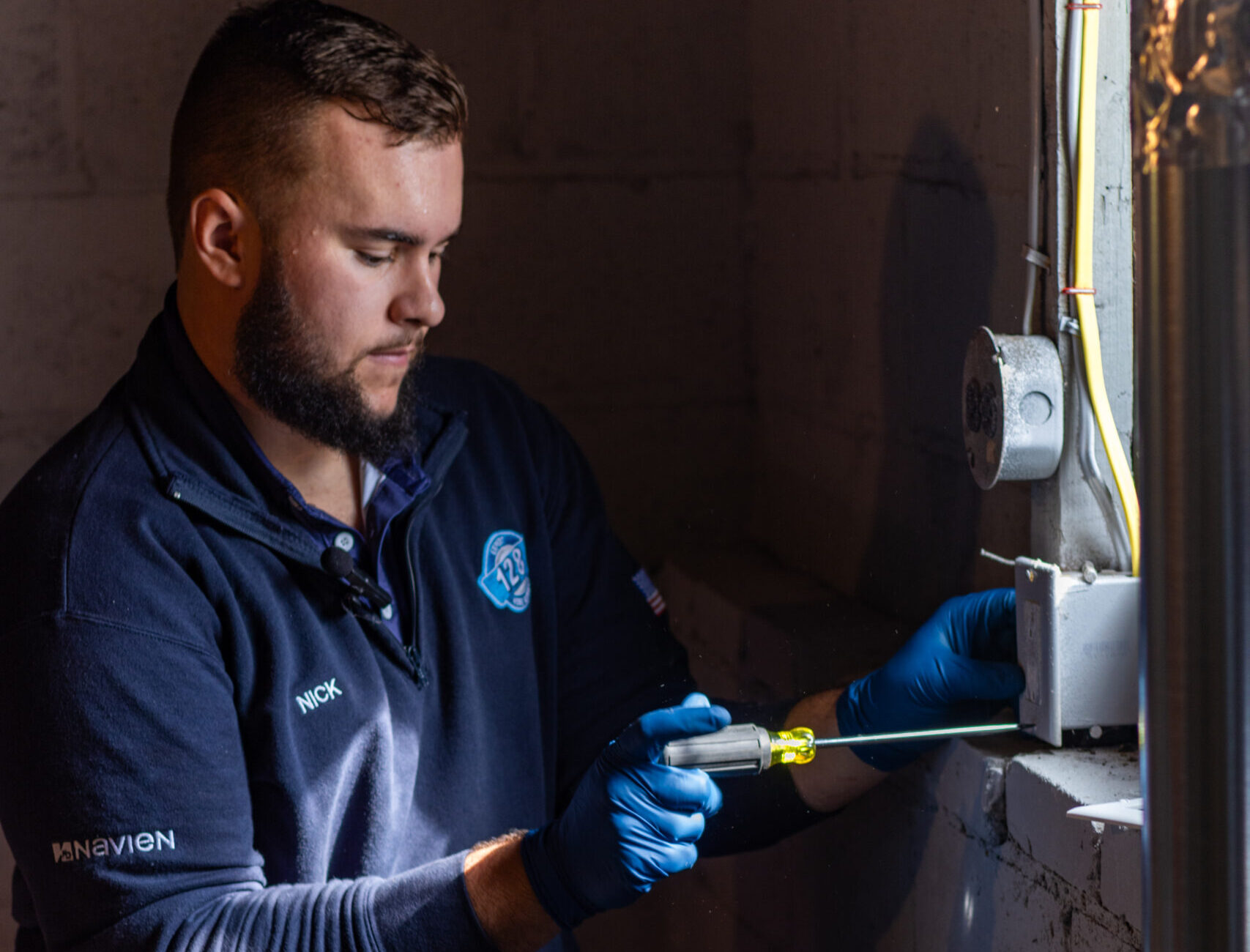A man wearing gloves and a tool belt uses a screwdriver to work on an electrical outlet in a basement or utility room.