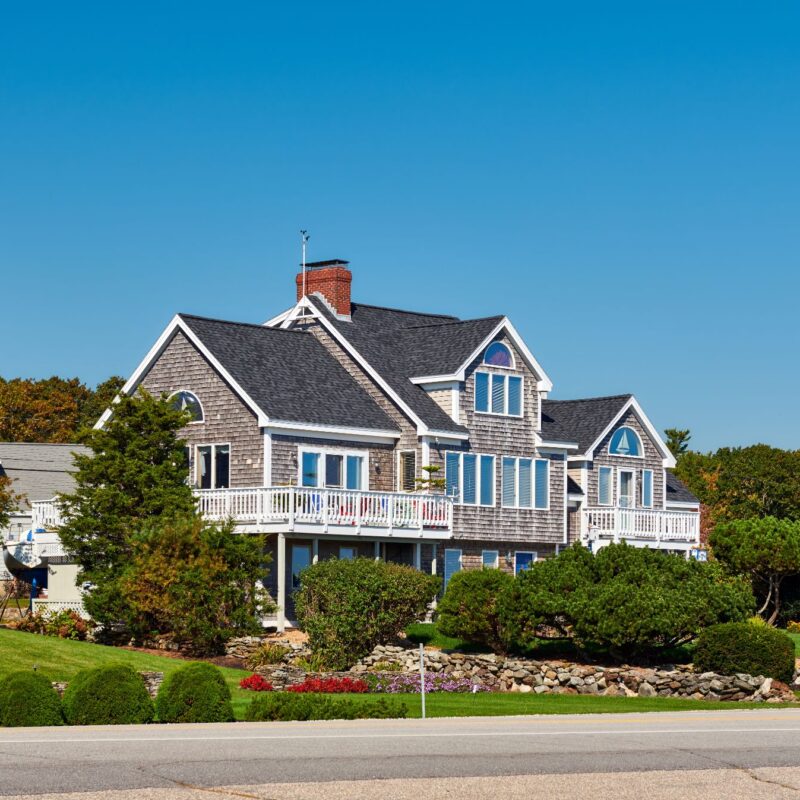 A large, gray shingle-style house with white trim and a wraparound porch in Chelmsford, surrounded by landscaped shrubs and a stone wall, under a clear blue sky.