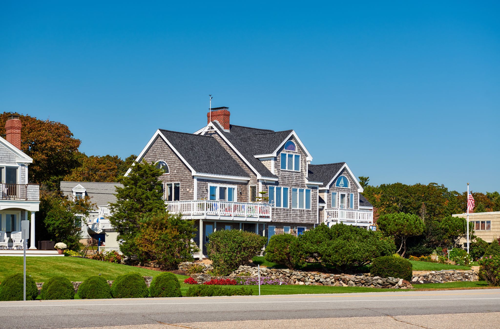 A large, gray shingle-style house with white trim and a wraparound porch in Chelmsford, surrounded by landscaped shrubs and a stone wall, under a clear blue sky.