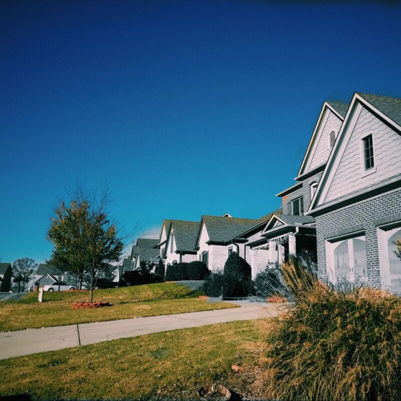 Row of modern suburban houses with driveways, neatly trimmed lawns, and bushes under a clear blue sky.