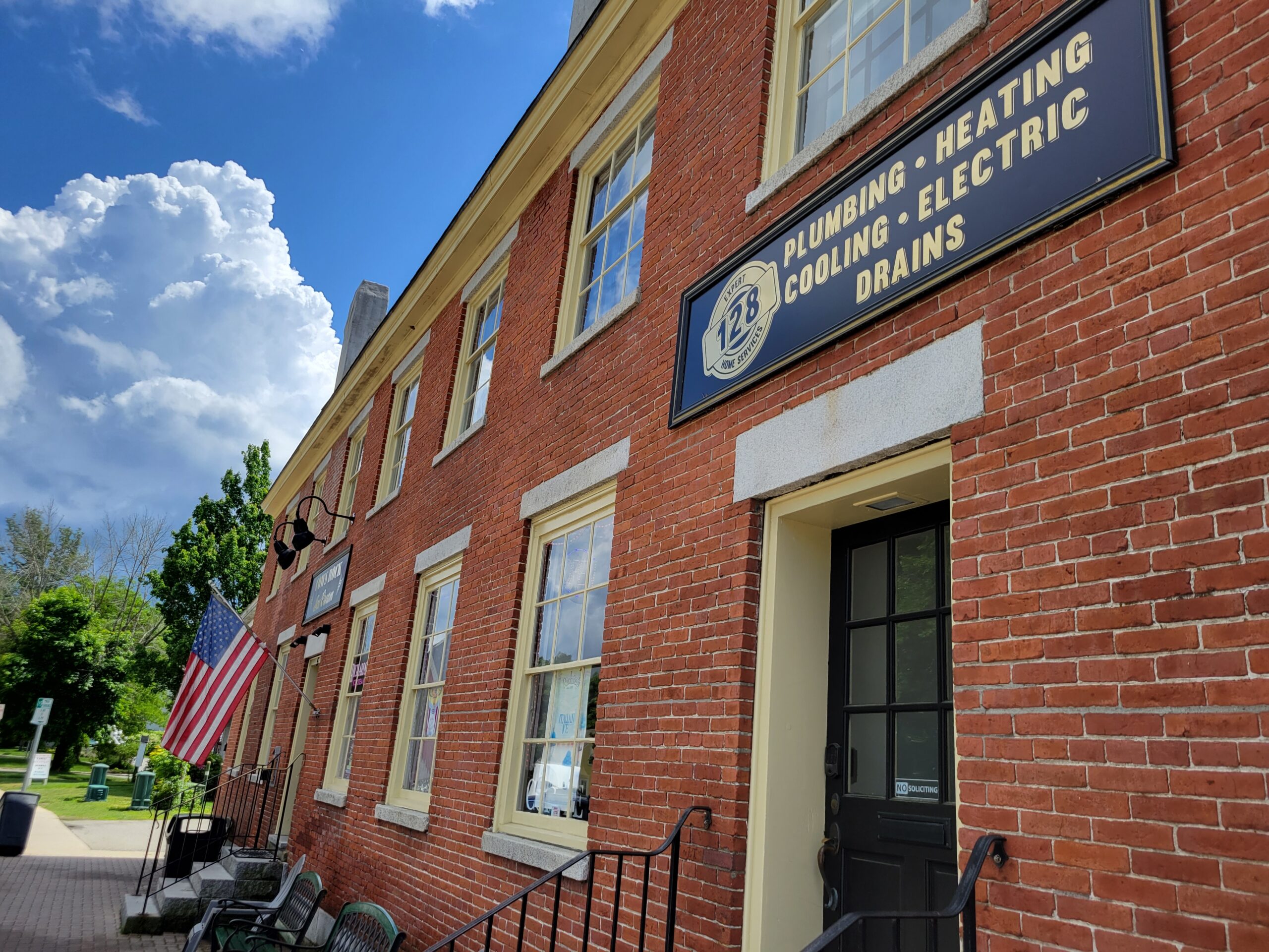 Red brick building with cream trim, American flag, and sign reading “Plumbing, Heating, Cooling, Electric, Drains” above a black door. Benches and greenery are visible outside—a local landmark for North Andover MA plumbing heating cooling & electric services.