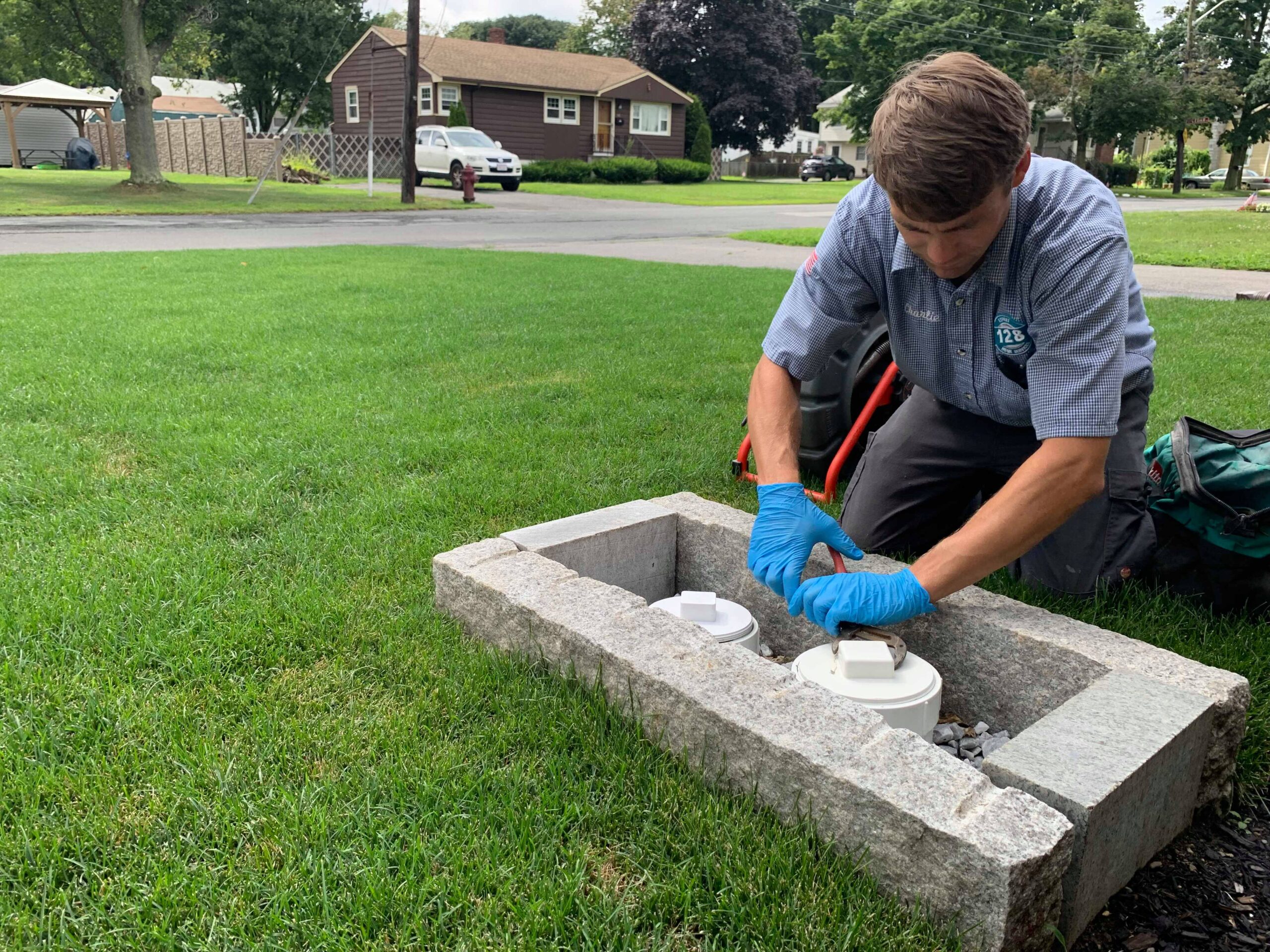 A person wearing gloves and a uniform installs or inspects equipment in a concrete enclosure on a grassy lawn in a residential neighborhood, likely as part of Winchester MA drain cleaning services.