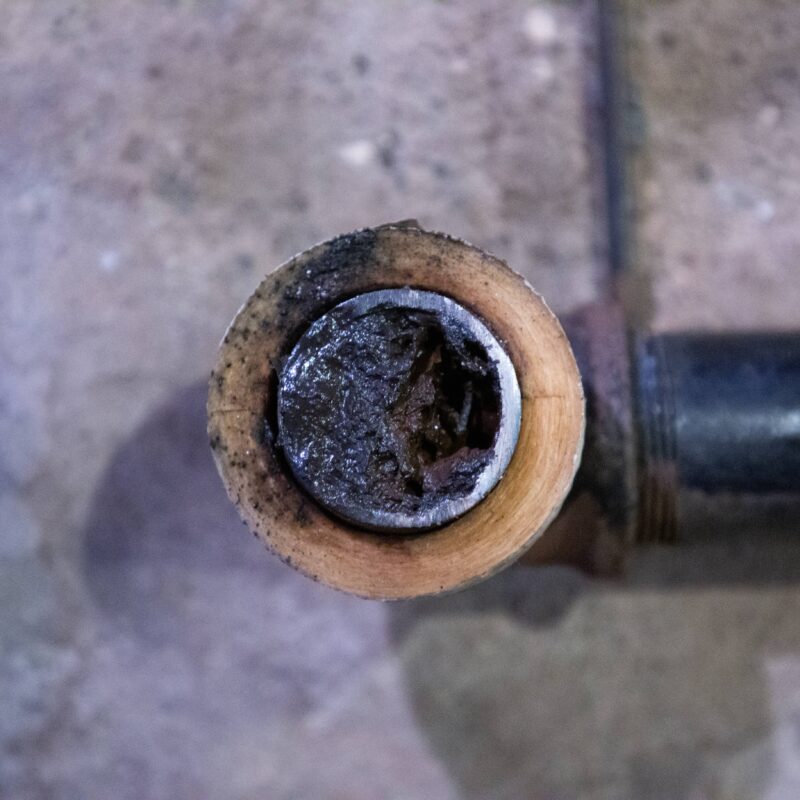 Close-up of the end of a metal pipe with a thick, dark buildup or blockage inside, resting on a concrete floor.
