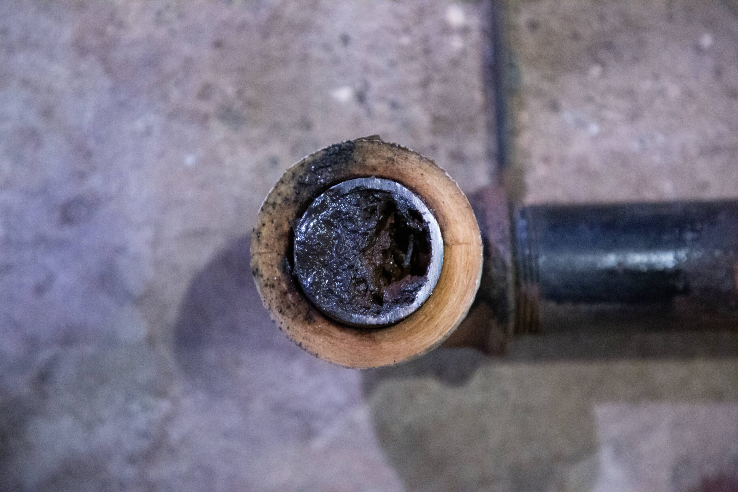 Close-up of the end of a metal pipe with a thick, dark buildup or blockage inside, resting on a concrete floor.