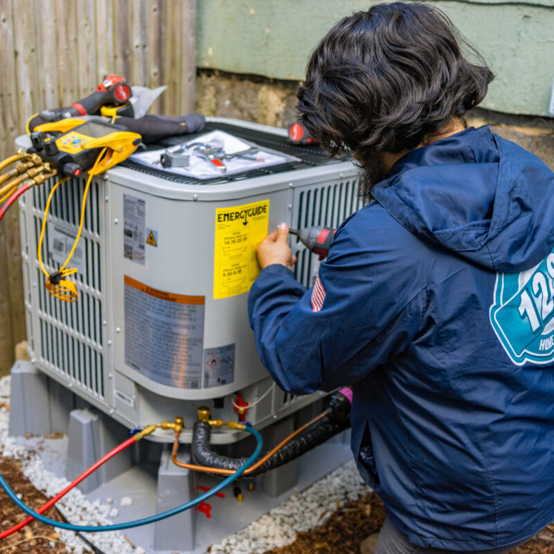 A technician in a blue jacket services an outdoor HVAC unit beside a house, using tools and connected gauges, while providing advice on how much does HVAC service cost in Haverhill MA.