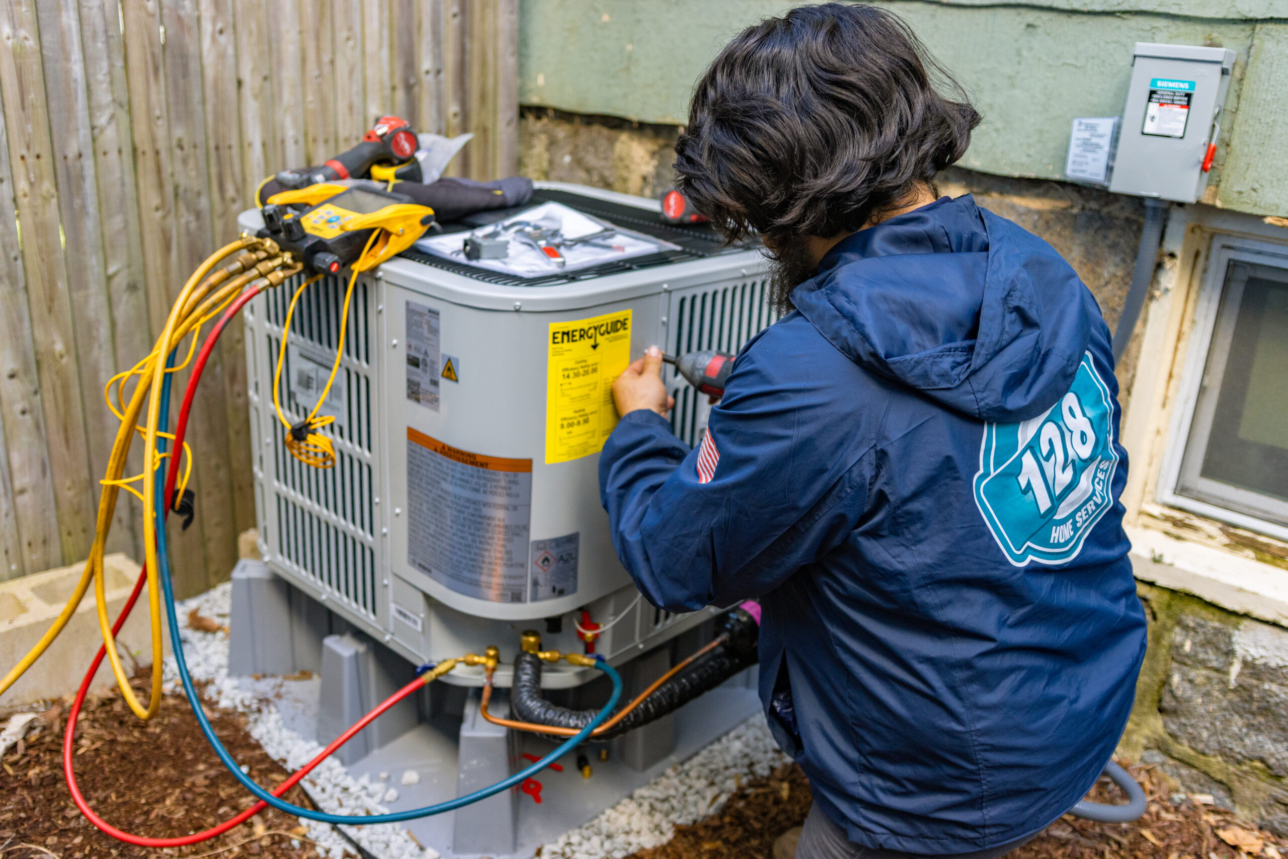 A technician in a blue jacket services an outdoor HVAC unit beside a house, using tools and connected gauges, while providing advice on how much does HVAC service cost in Haverhill MA.