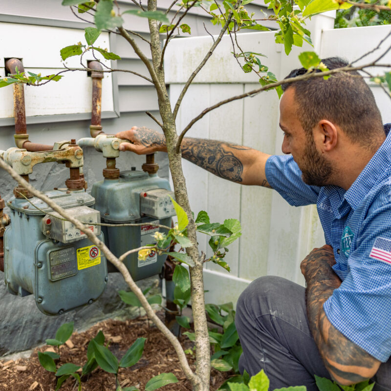 A man in a blue shirt inspects two outdoor gas meters attached to the side of a house, surrounded by plants and a white fence—something you might need to do if you’re learning how to find a licensed plumber in Wakefield, MA.