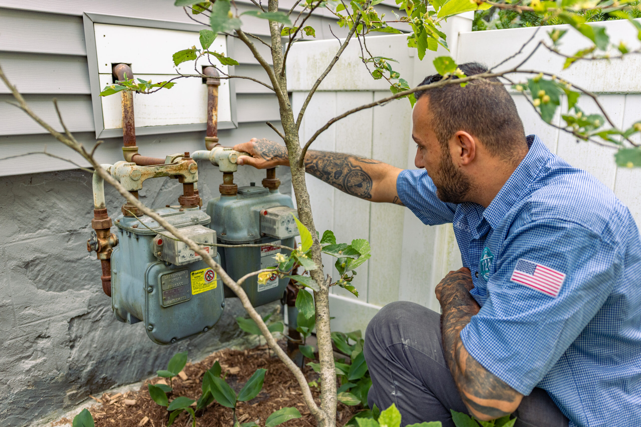 A man in a blue shirt inspects two outdoor gas meters attached to the side of a house, surrounded by plants and a white fence—something you might need to do if you’re learning how to find a licensed plumber in Wakefield, MA.