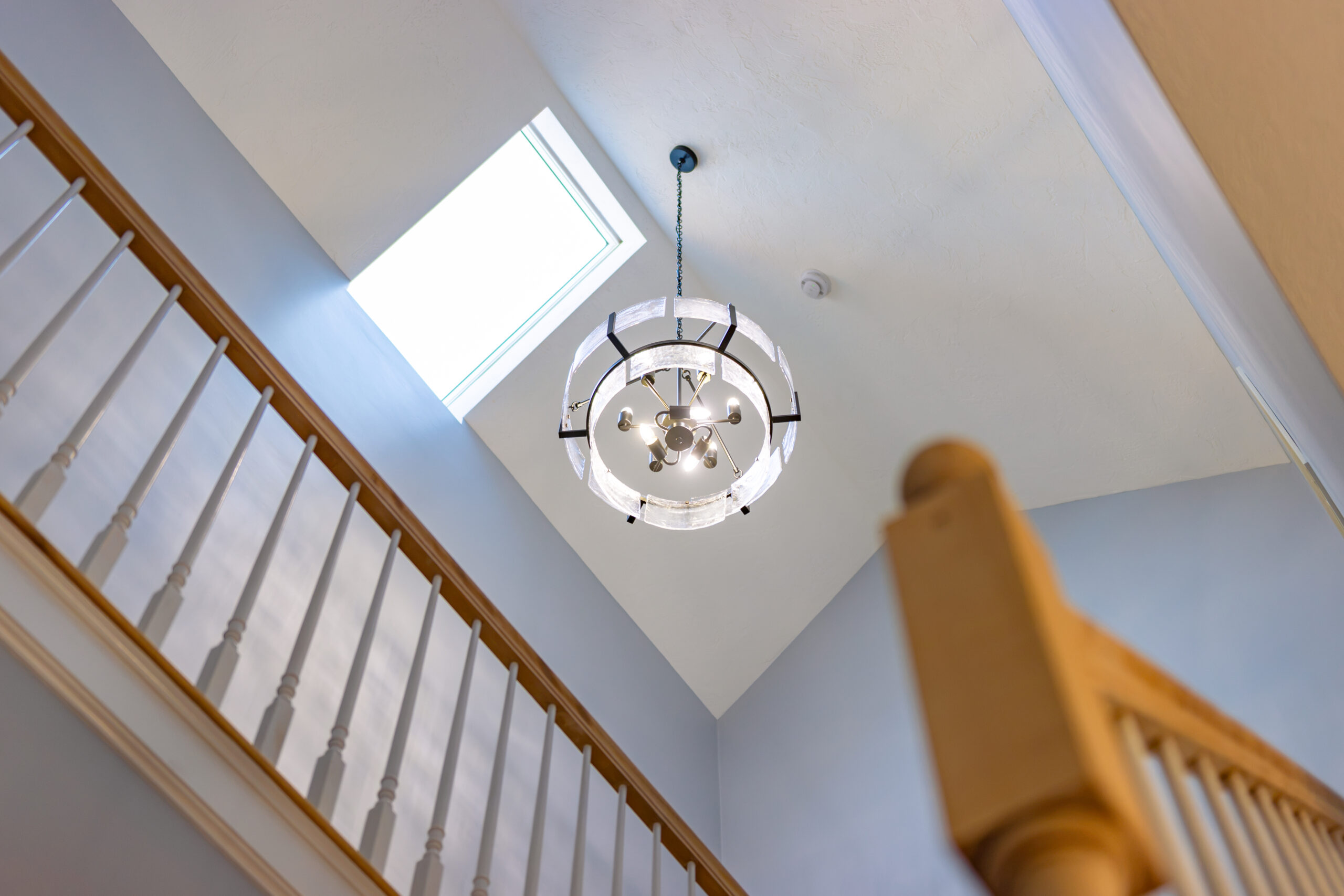 A multi-tiered, spherical chandelier hangs from a vaulted ceiling near a skylight above a staircase with wooden railings.