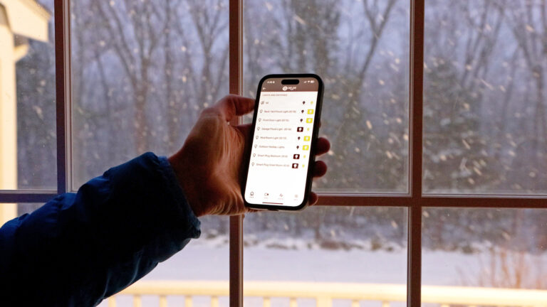 A hand holds a smartphone displaying a smart home app screen listing light and switch controls, with a snowy outdoor scene visible through a window in the background.