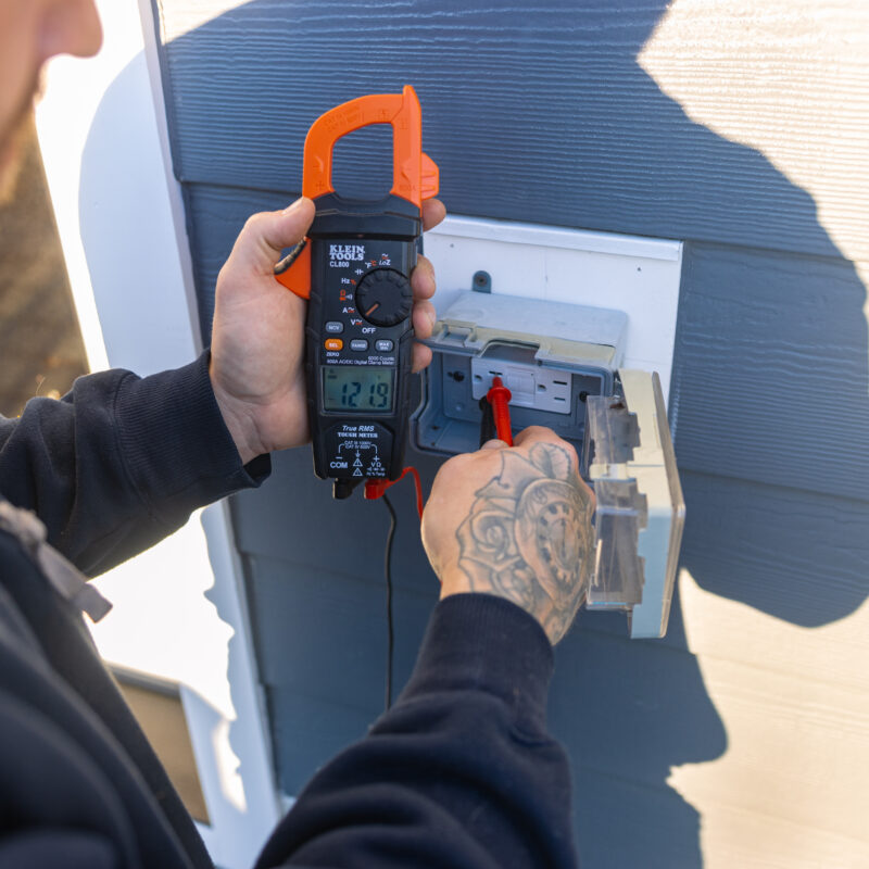 Person measuring voltage with a digital clamp meter on an outdoor electrical panel mounted on a blue wall.
