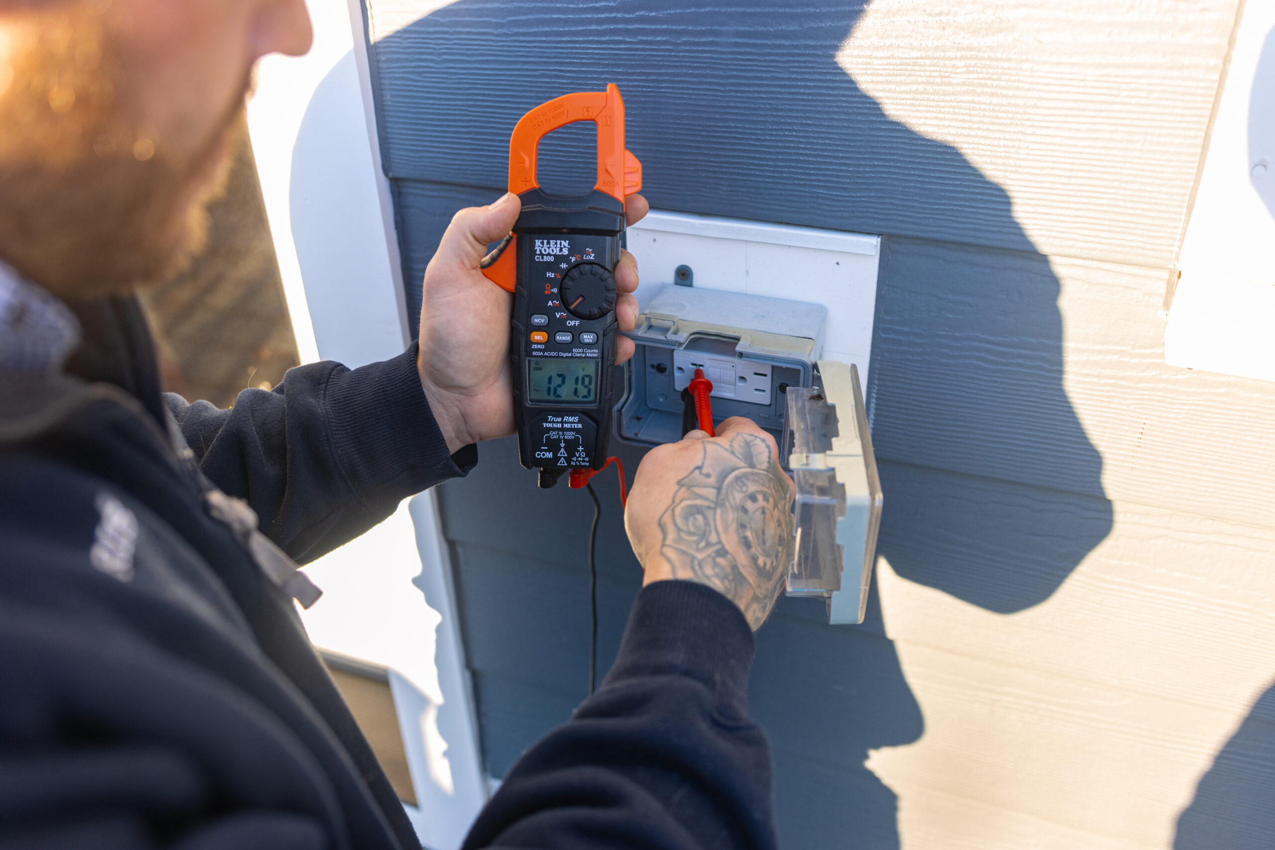 Person measuring voltage with a digital clamp meter on an outdoor electrical panel mounted on a blue wall.